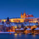 Prague Castle and Charles Bridge illuminated at dusk reflecting on the Vltava River with snow-covered buildings